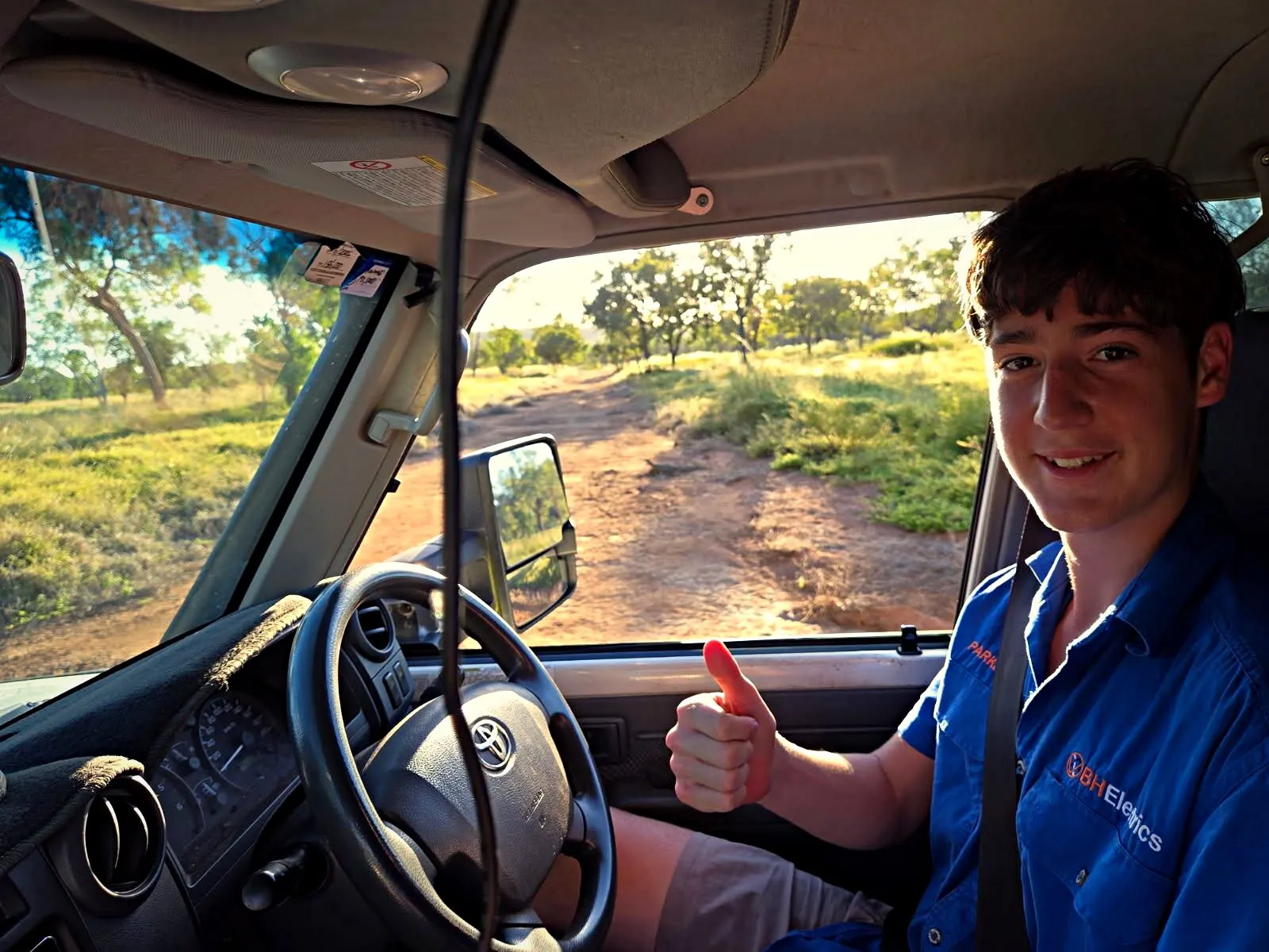 A BH Electrics apprentice at the wheel of the ute on a red dirt road, giving a thumbs up on the way to a remote job