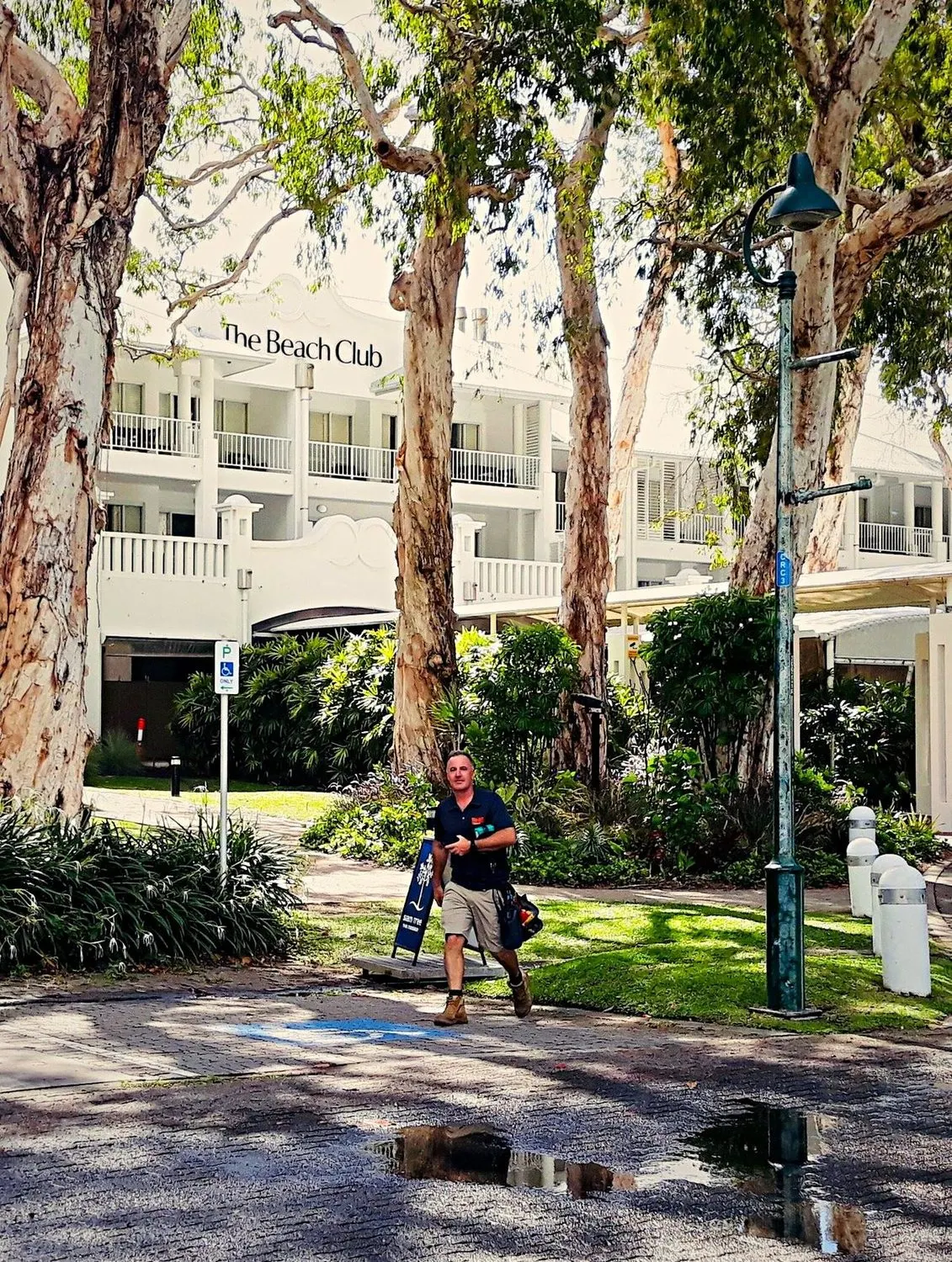 A BH Electrics electrician walking in to a job at The Beach Club, Palm Cove, tool bag in hand, under the paperbarks