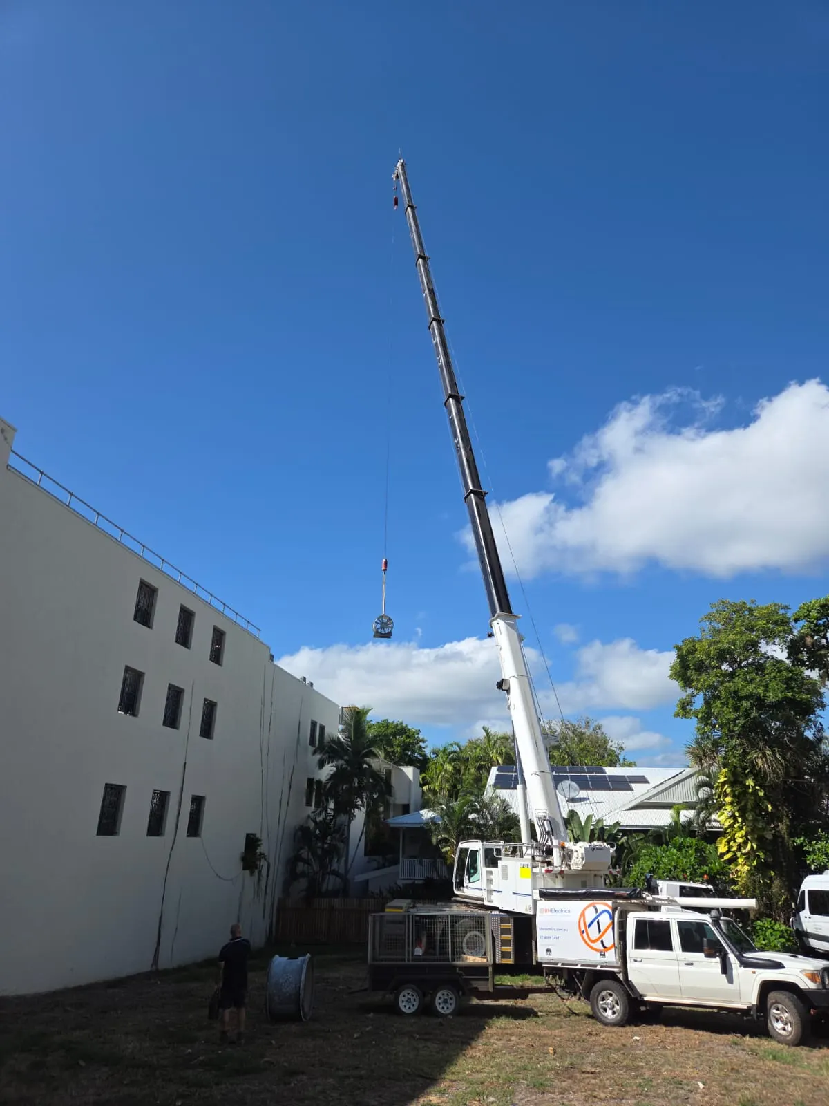 A crane lifting plant onto the roof of a Port Douglas building, with the BH Electrics ute and cable drum trailer on the ground below