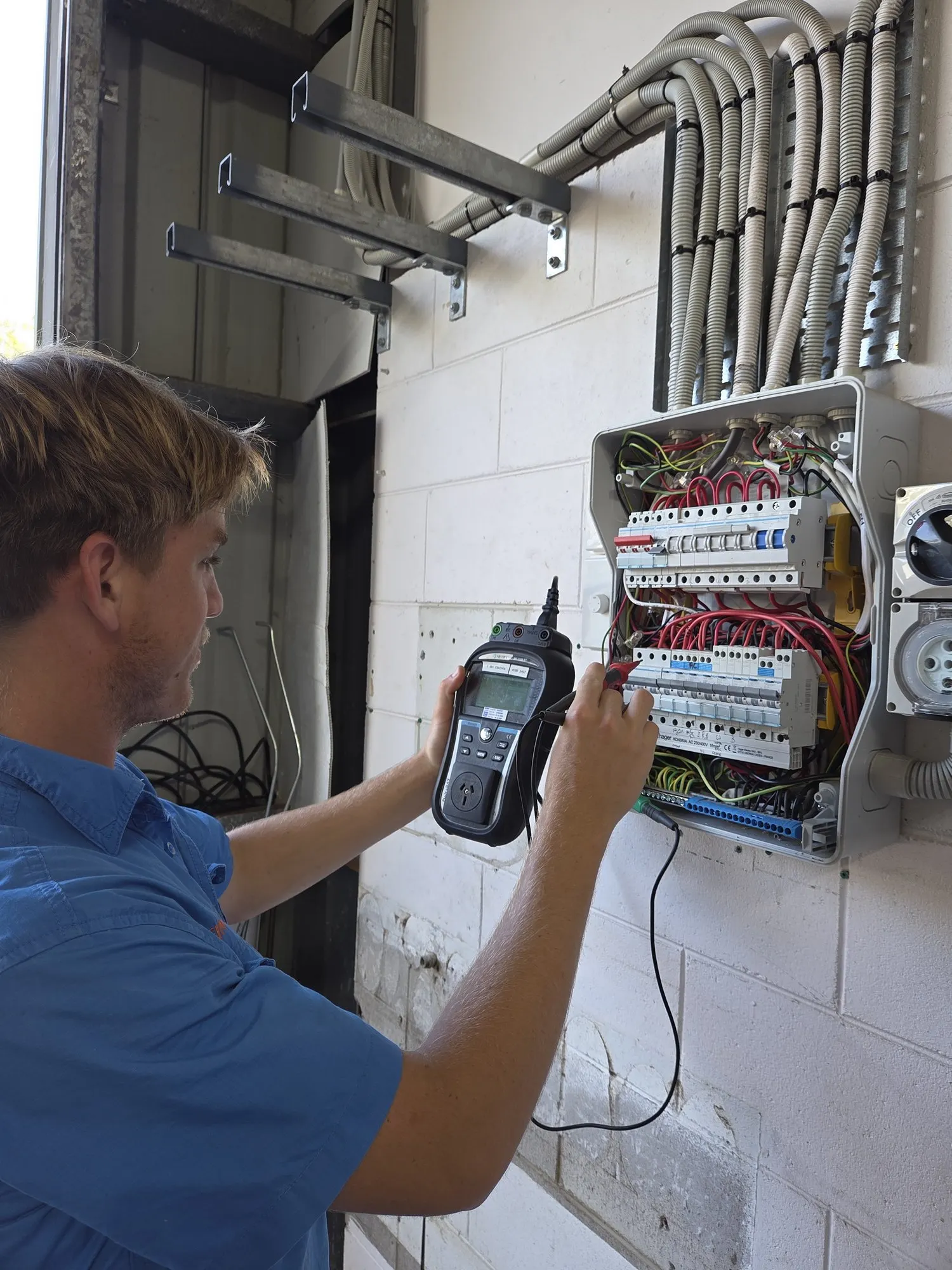 A BH Electrics electrician testing a safety switch at an open switchboard with a multifunction tester