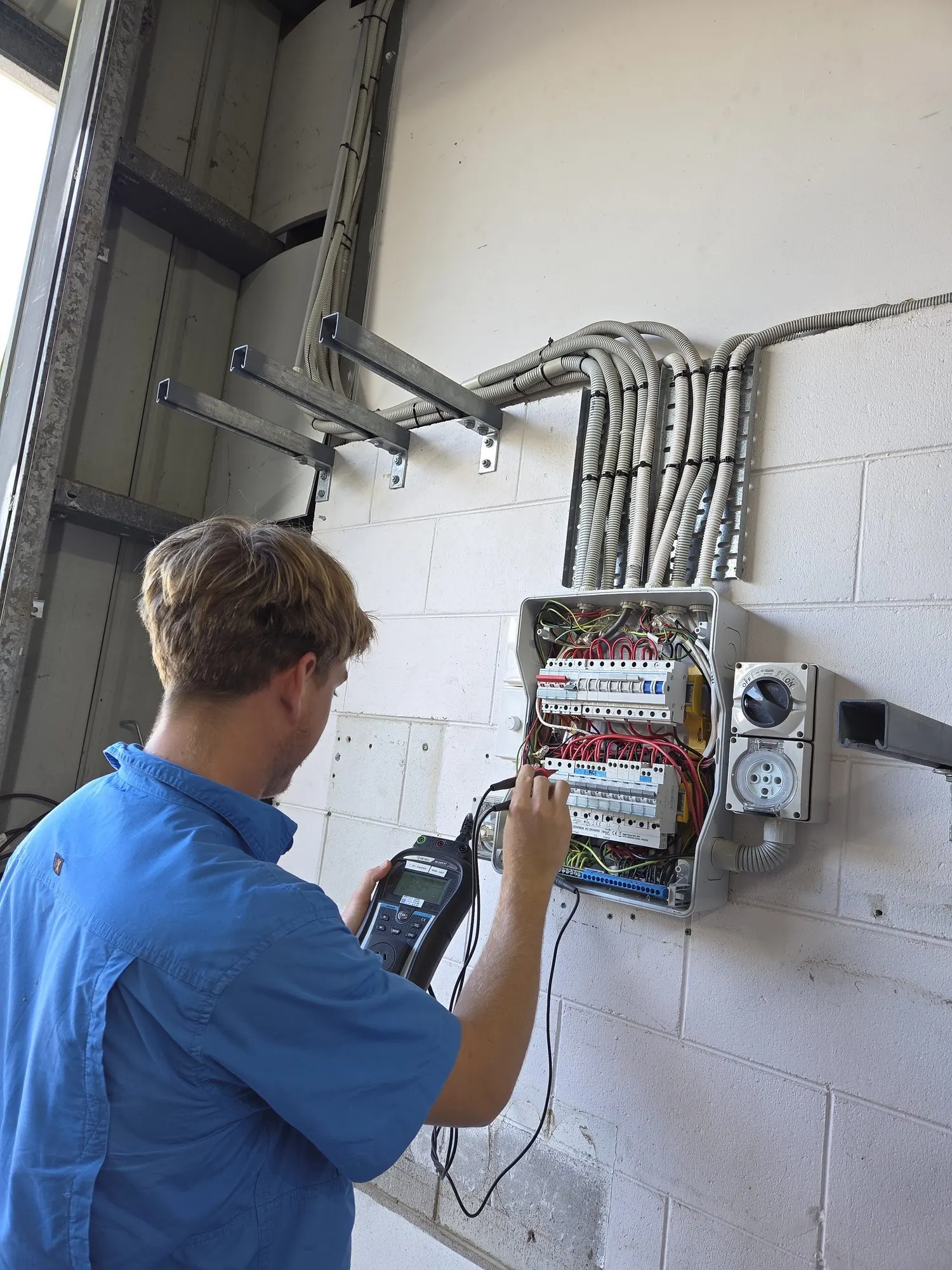 An electrician testing safety switches at a commercial switchboard, with the conduit run and isolator alongside