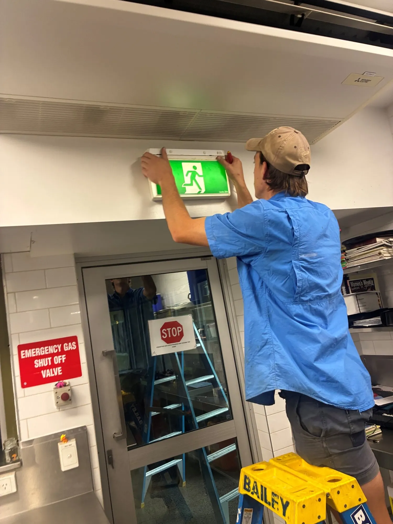 A BH Electrics electrician on a platform ladder working on an illuminated exit sign above a commercial kitchen door