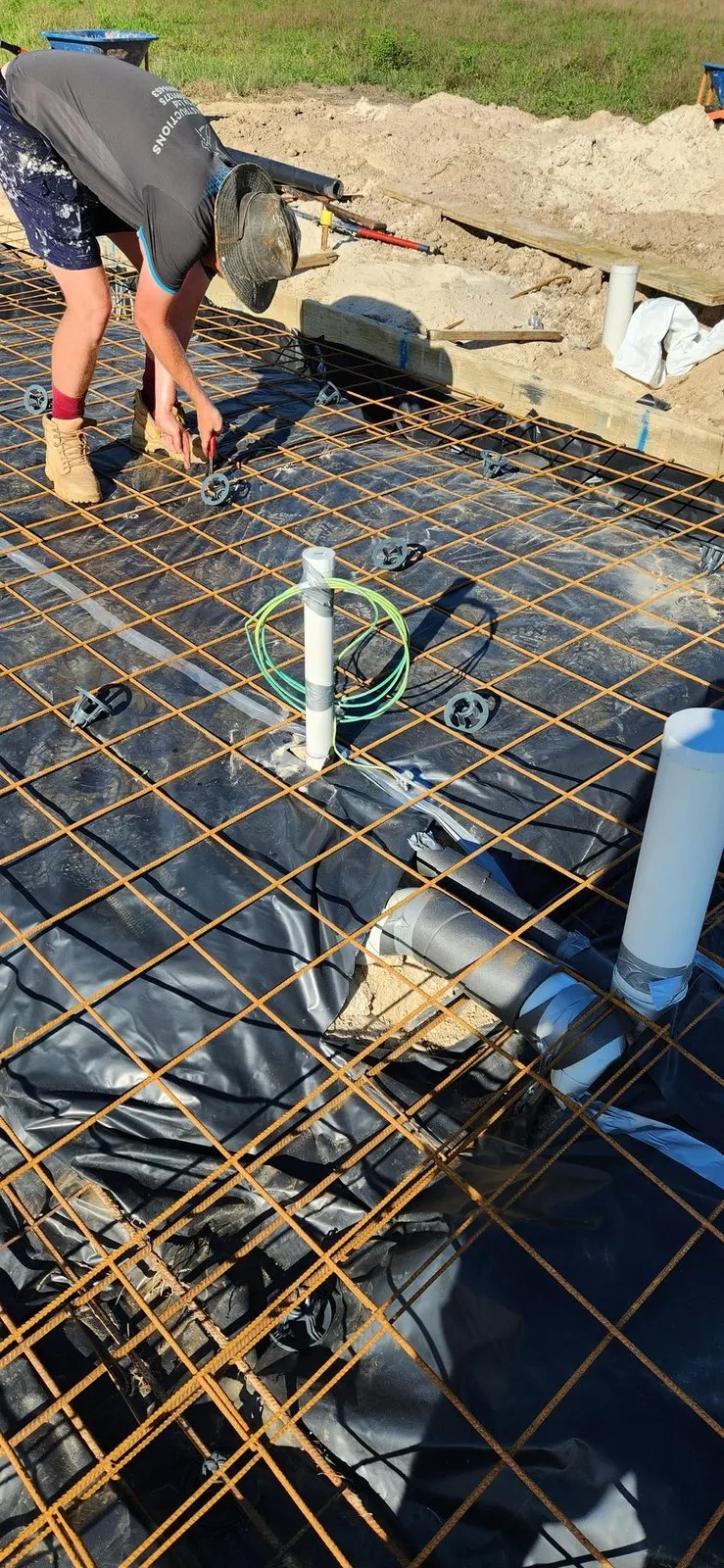 Conduit and earth cable set through the membrane in a slab before the pour, with a builder tying off the reinforcing