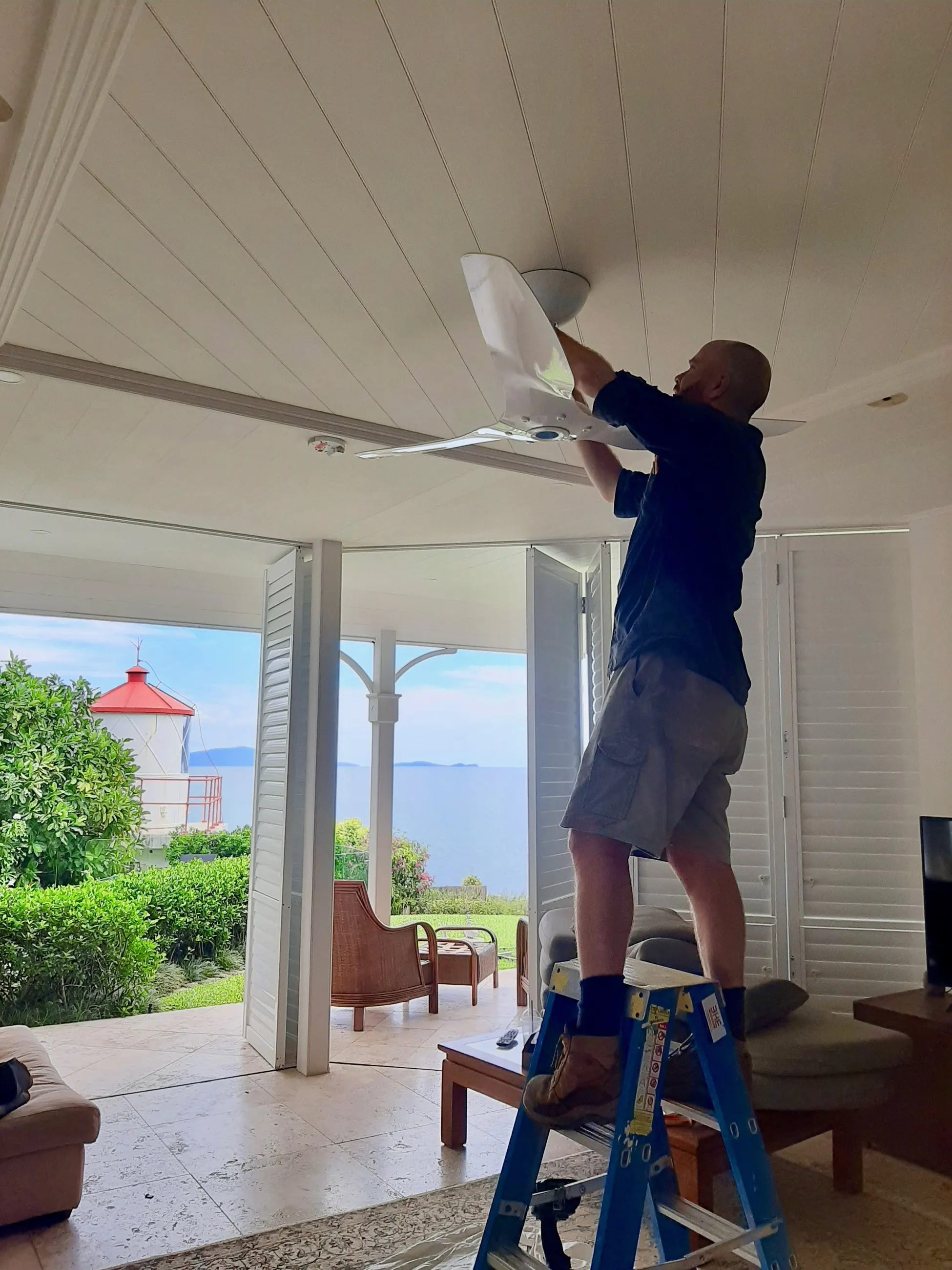 A BH Electrics electrician on a step ladder fitting a ceiling fan on an open verandah, water and a red roof beyond