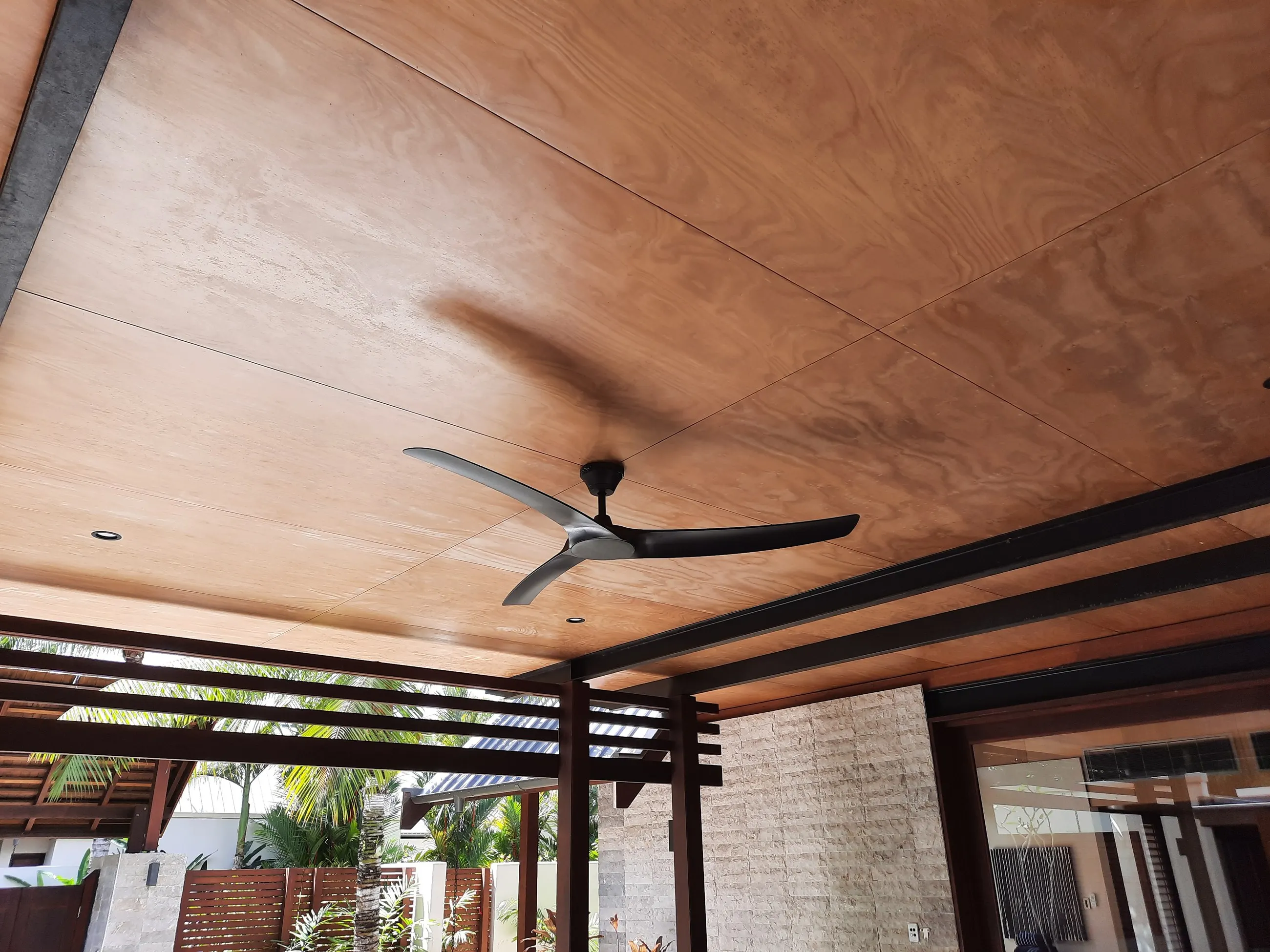 A dark ceiling fan fitted to a timber-lined patio ceiling, pergola and garden beyond