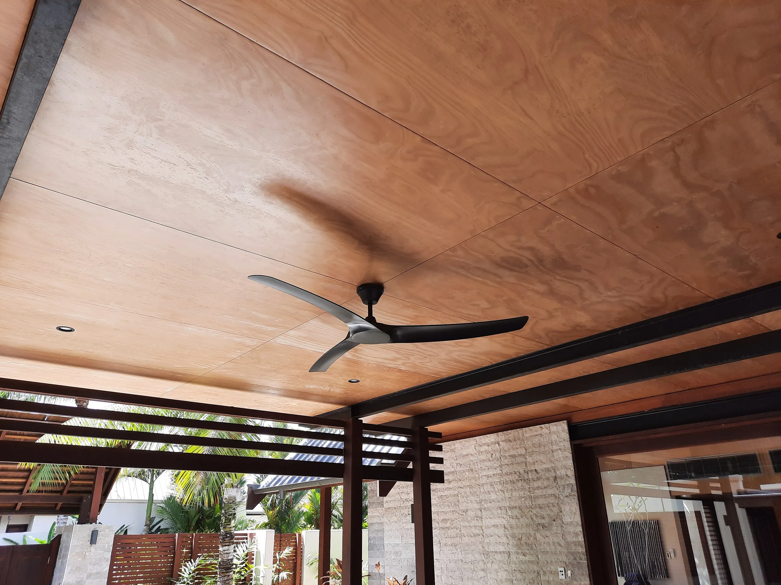 A dark ceiling fan fitted to a timber-lined patio ceiling, pergola and garden beyond