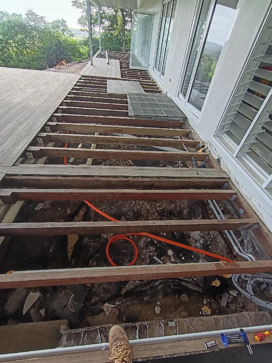 Exposed deck joists during a rebuild, with new electrical conduit and cable run alongside, glass doors and hinterland beyond