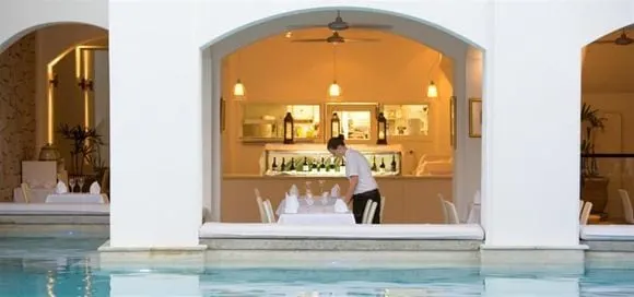 Looking through an arch across the pool into the lit dining room at Peppers Beach Club, pendant lights and ceiling fans inside