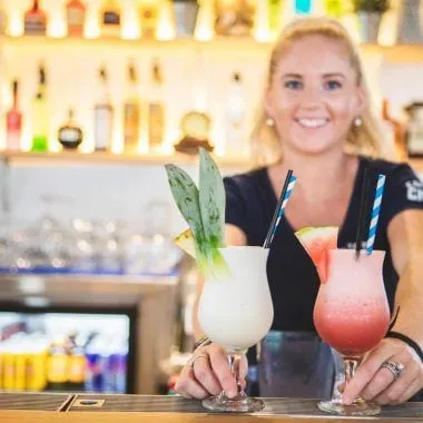 A Court House Hotel bartender holding two cocktails across the bar, the lit back bar shelves behind her