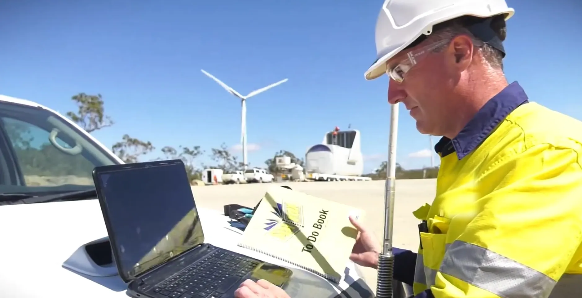 Brad Sims in a hard hat and hi-vis working at a laptop on the bonnet of a ute at Mount Emerald, a turbine and a nacelle behind him