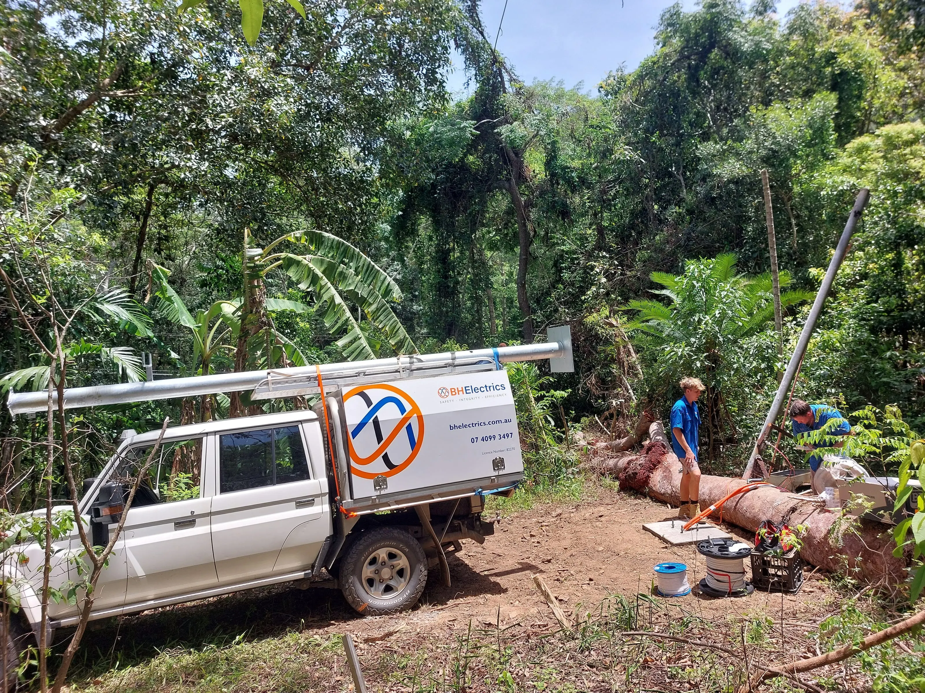 A BH Electrics ute at a property at Mowbray after Cyclone Jasper, a galvanised pole on the roof racks and two electricians at the new footing beside the fallen tree